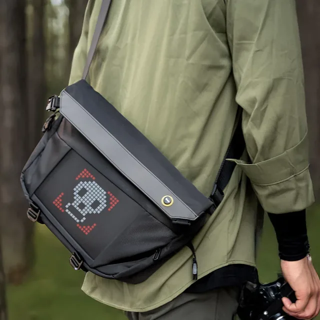 Человек с камерой несёт чёрную Pixoo Sling Bag-V через плечо, на экране светится пиксельный рисунок