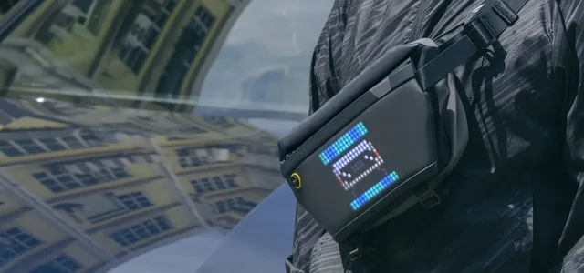 Divoom Pixoo Sling Bag 4 надета через плечо: на пиксельном экране синяя анимация, сумка снята крупным планом на фоне стеклянного фасада