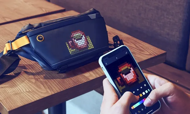 Divoom Pixoo Sling Bag 4 на столе: на экране сумки пиксельный персонаж, рядом смартфон с редактором пиксель-арта в приложении Divoom
