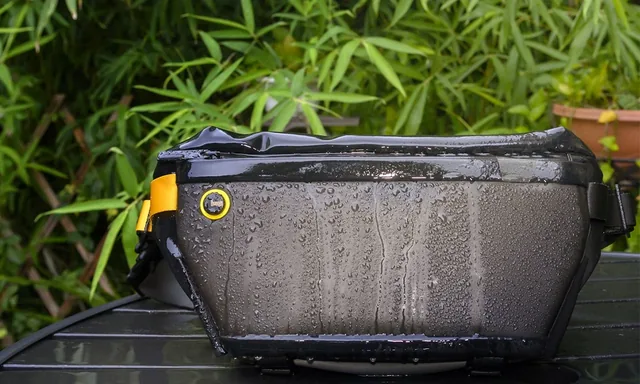 Divoom Pixoo Sling Bag 4 в каплях воды после дождя: водоотталкивающая эко-ткань корпуса и жёлтая стропа, фон — зелёные растения